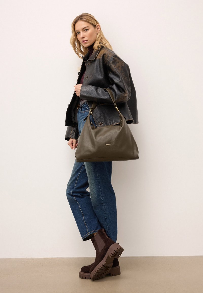 Olíva zöld bőr kézitáska textúrált felülettel, arany színű fém kiegészítőkkel és laza fazonnal. Sötét dzsekivel és kék farmernadrággal viselve.