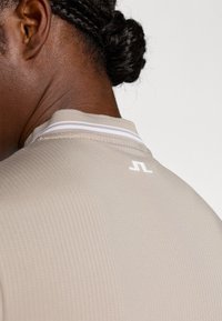 Maglietta sportiva beige con un tessuto testurizzato e una striscia bianca accentata sul colletto. Presenta un piccolo logo bianco sul retro del collo.