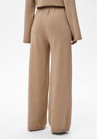 Bronzen wijde broekspijpen gemaakt van glinsterende stof. Voorzien van een elastische tailleband en geribbelde boorden. Zachte textuur met een ontspannen pasvorm.