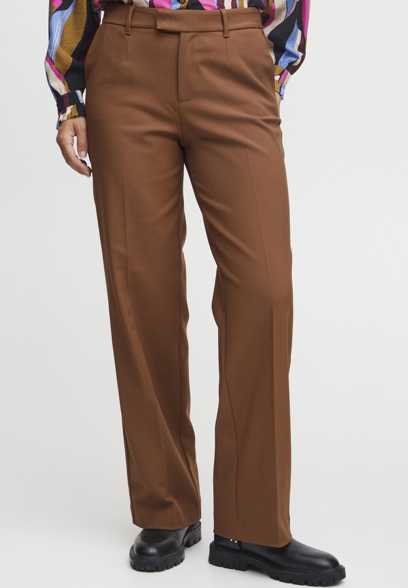Personne portant un pantalon marron à jambes droites, un chemisier rayé multicolore et des chaussures noires à semelle épaisse, debout sur un fond uni.