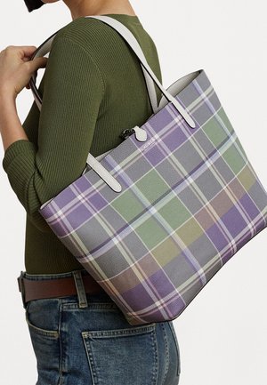Persona che indossa una maglia verde a maniche lunghe a coste e jeans, che tiene una grande borsa a tracolla a quadri con sfumature di viola, verde e beige.