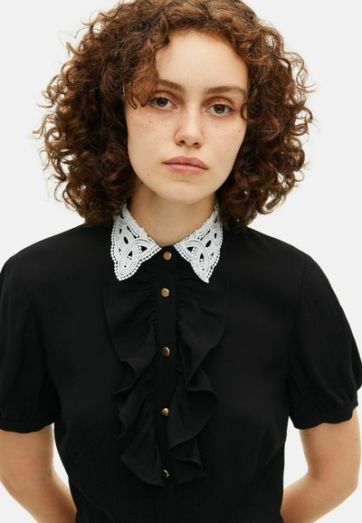 Bluză neagră cu un guler Peter Pan din dantelă albă, cu închidere prin nasturi și detalii volănaș. Mâneci scurte tip puff. Textură de stofă netedă.