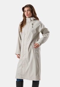 Femme portant un long imperméable gris clair avec des boutons et des poignets ajustables, debout avec une main dans la poche devant un fond uni.