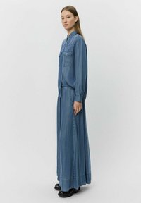 Vaaleansininen denim-paita ja pitkä hame, jossa on kaksi rintap pocketia ja kerätty vyötärö. Mallilla on päällään mustat kengät. Yksinkertainen tausta.