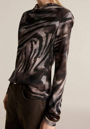 Femme portant un haut à manches longues à col haut avec un imprimé animal abstrait noir et gris, assorti à un pantalon marron foncé et de grands bracelets au poignet.