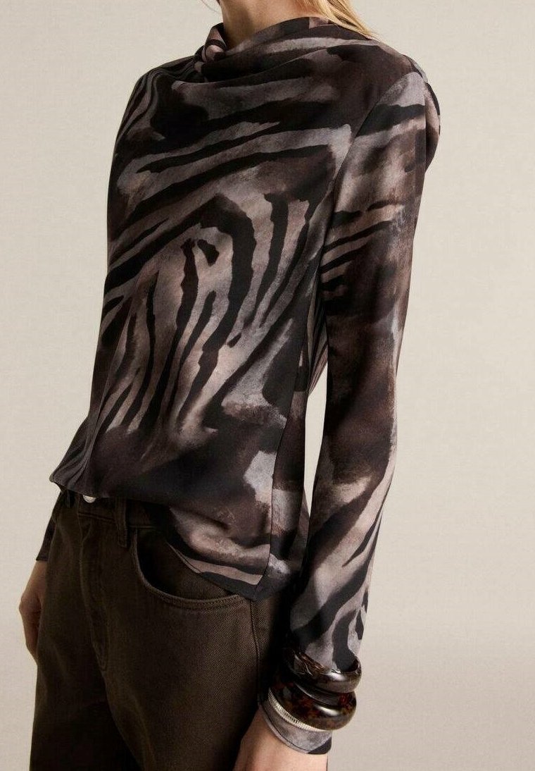 Femme portant un haut à manches longues à col haut avec un imprimé animal abstrait noir et gris, assorti à un pantalon marron foncé et de grands bracelets au poignet.
