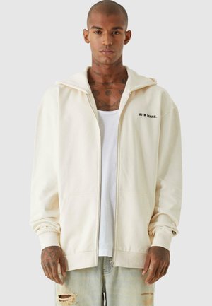 Cremefarbener Reißverschluss-Hoodie mit weicher Textur, Seitentaschen und gesticktem Logo auf der linken Brust. Über einem weißen T-Shirt getragen und kombiniert mit zerrissenen Jeans.