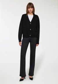 Cardigan nero a maglia con scollo a V, cinque bottoni e due tasche frontali. Abbinato a pantaloni a righe bianchi e neri e tacchi a punta neri.