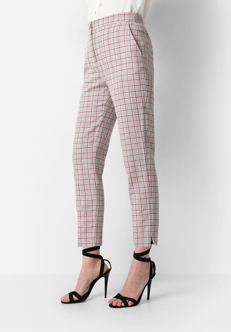 Pantalon à carreaux gris clair avec des lignes rouges et noires. Coupe ajustée avec poches latérales et fentes aux chevilles, porté avec des sandales noires à talons.