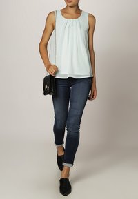Top senza maniche di un blu chiaro con pieghe, abbinato a jeans di denim scuri e scarpe nere. La modella porta una piccola borsa a tracolla nera con una tracolla a catena.