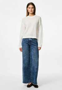 Biały dzianinowy sweter o luźnym kroju i falbaniastym dół, zestawiony z szerokimi ciemnoniebieskimi dżinsami. Modelka nosi czarne buty typu slip-on.