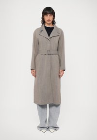 Světle šedý dlouhý kabát s límcem, páskem v pase a texturovanou látkou; v kombinaci se světle šedými kalhotami s širokými nohavicemi a špičatými bílými botami.