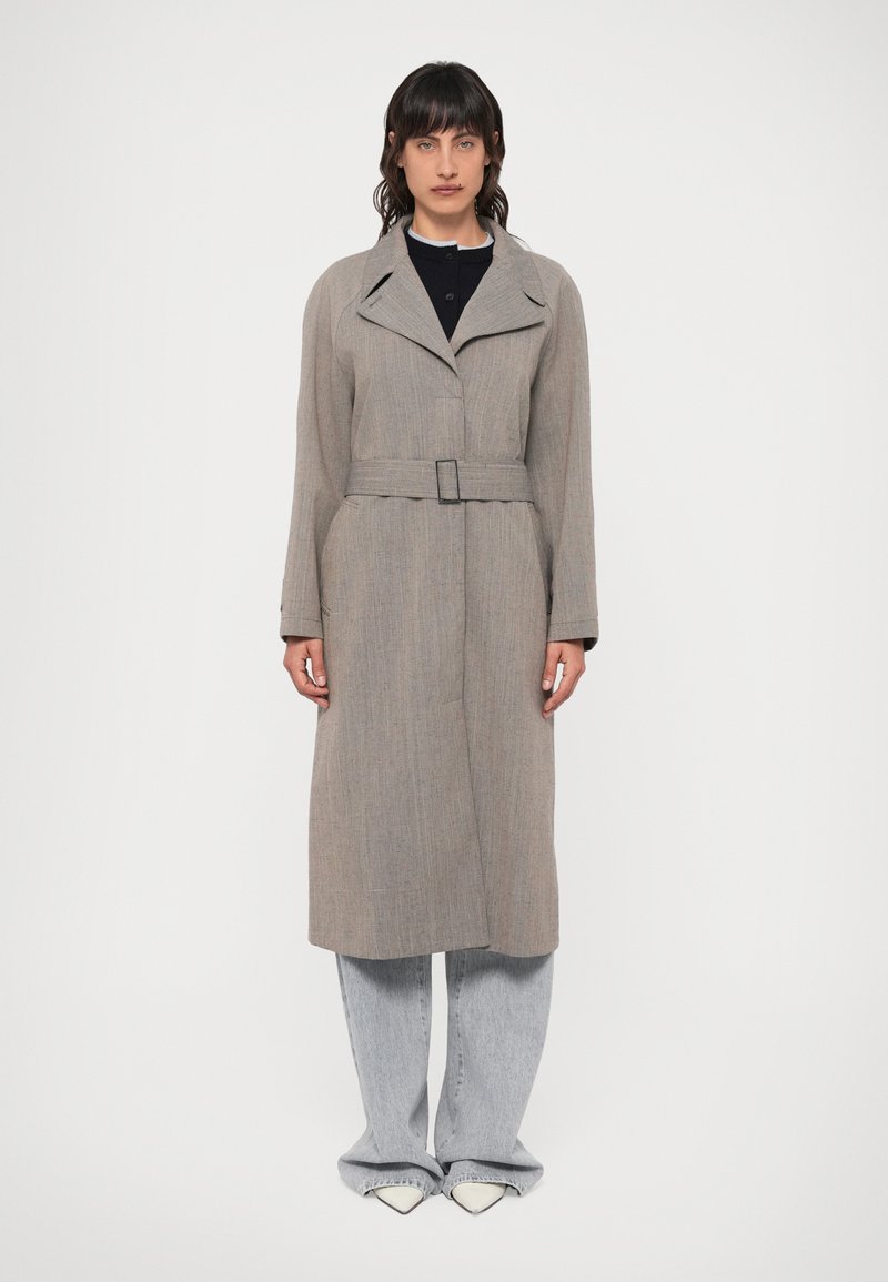 Světle šedý dlouhý kabát s límcem, páskem v pase a texturovanou látkou; v kombinaci se světle šedými kalhotami s širokými nohavicemi a špičatými bílými botami.