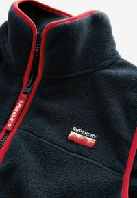 Fleece'owa kurtka w kolorze granatowym z czerwonymi akcentami. Wysoki kołnierz i zamek błyskawiczny z naszywką markową. Miękka tekstura z kontrastującymi detalami szycia.