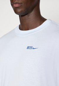 Hvid bomuldstrøje med blåt broderet "INTER" logo og Nike swoosh. Rund hals, korte ærmer, blød tekstur, afslappet pasform.