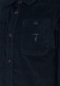 Chemise en velours côtelé bleu marine avec un devant à boutons, deux poches poitrine et un logo brodé discret sur la poche gauche.