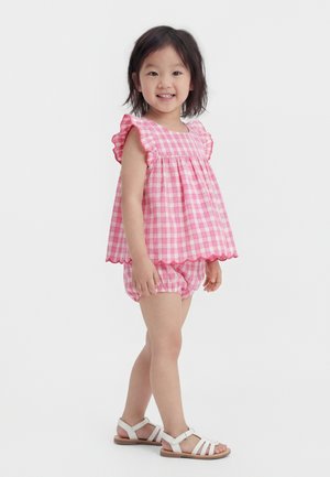 Šypsosi jauna mergina stovinti, dėvinti rožinį languotą be rankovių viršų su raukinukais ir derančius šortus bei baltus sandalus baltame fone.