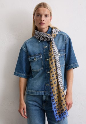 Chemise en denim à manches courtes avec deux poches poitrine, portée avec une écharpe à motifs en jaune, blanc et bleu, présentant des motifs géométriques et des bords frangés.