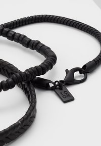 Svart lær lanyard med teksturerte, sylinderformede perler og en flettet del. Inkluderer en svart klips og en liten merkelapp med "ICON" preget.