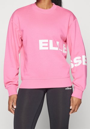 Person trägt ein leuchtend pinkes Sweatshirt mit weißem "ellesse"-Schriftzug und schwarze Leggings mit einem kleinen weißen "ellesse"-Logo.