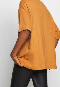 Tričko s krátkými rukávy v hořčicové barvě, vyrobené z měkké bavlny. Má volný střih s zaobleným lemem a mírnými bočními rozparky.