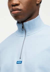 Κοντινό πλάνο ενός ατόμου που φοράει ανοιχτό μπλε ζακέτα με φερμουάρ, ψηλό γιακά και μικρό μπλε λογότυπο «HUGO» στο στήθος.