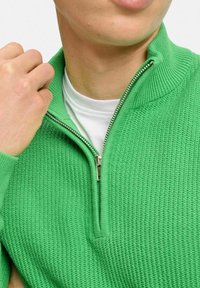 Grön zip-up tröja med en texturerad stickad design. Har en hög krage, silverdragkedja och en lager vit t-shirt under.