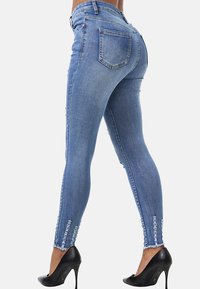 Jeans de denim azul ajustados con cintura alta, detalles desgastados en el dobladillo y dos bolsillos traseros. Combinados con zapatos de tacón alto negros.