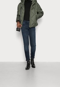 Ciemnozielona kurtka puchowa z kapturem, zestawiona z ciemnoniebieskimi dżinsami i czarnymi botkami za kostkę. Prosty fason z pikowaną teksturą.