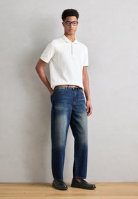 Polo bianco, jeans blu scuro con una texture sbiadita e scarpe slip-on nere. Il modello indossa occhiali e mantiene una postura rilassata.