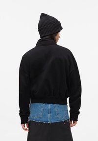 Sudadera negra de gran tamaño con cuello alto, puños acanalados y un dobladillo corto. Combinada con una camisa de mezclilla azul debajo.