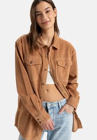Bruine corduroy overhemd met zichtbare textuur, een kraag, knoopsluitingen en twee voorzakken, gecombineerd met een witte top en lichtblauwe jeans.