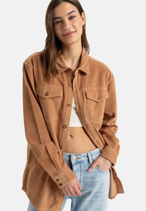 Bruine corduroy overhemd met zichtbare textuur, een kraag, knoopsluitingen en twee voorzakken, gecombineerd met een witte top en lichtblauwe jeans.