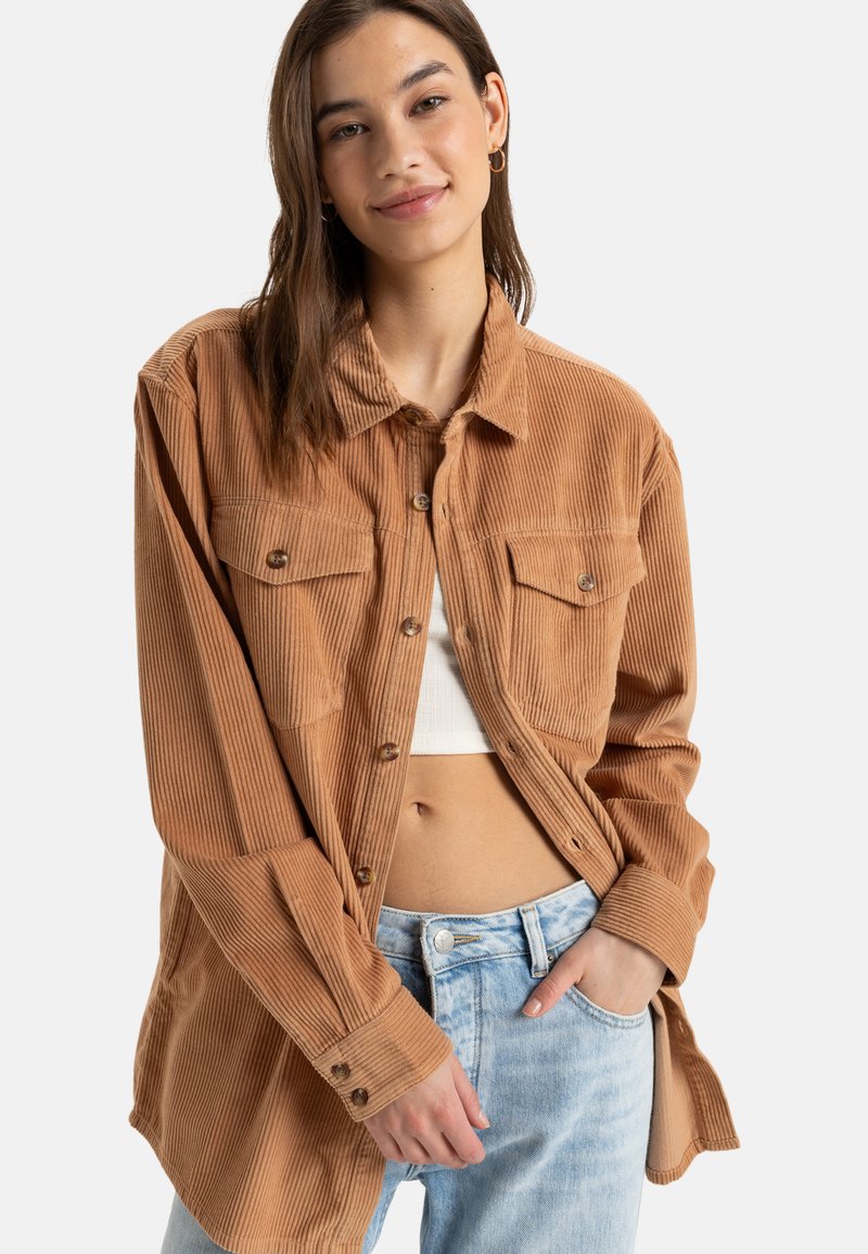 Bruine corduroy overhemd met zichtbare textuur, een kraag, knoopsluitingen en twee voorzakken, gecombineerd met een witte top en lichtblauwe jeans.