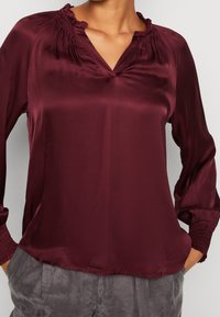 Burgunderfarbene Satinbluse mit V-Ausschnitt, gerafftem Kragen und elastischen Bündchen. Glatte Textur, lockere Passform und fällt knapp unter die Taille.