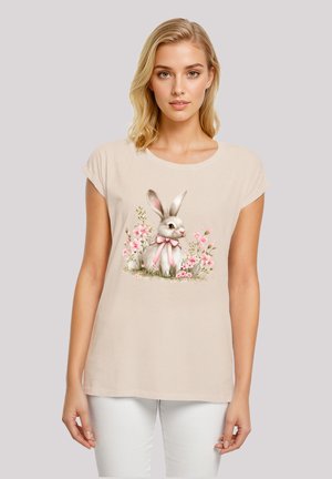 Beige T-Shirt mit kurzen Ärmeln, featuring ein gedrucktes Kaninchen mit rosa Schleife, umgeben von rosa Blumen, auf einem weichen, glatten Stoff.