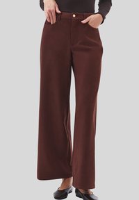 Persoon in bruin, hoge taille, wijd-pijped pantalons met voorzakken en zwarte platte schoenen, staand tegen een effen achtergrond.