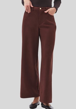 Persona che indossa pantaloni marroni a vita alta e a gamba larga con tasche frontali e scarpe basse nere, in piedi contro uno sfondo neutro.