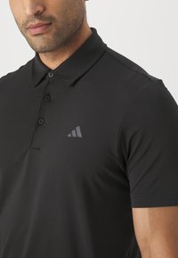 Polo preto com gola, mangas curtas, abertura com botões e um detalhe de logotipo cinza escuro subtil no lado esquerdo do peito. Textura lisa.