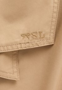 Beige textil med en strukturerad finish; har ett broderat "SL" och en dekorativ rosett nära sömmen.
