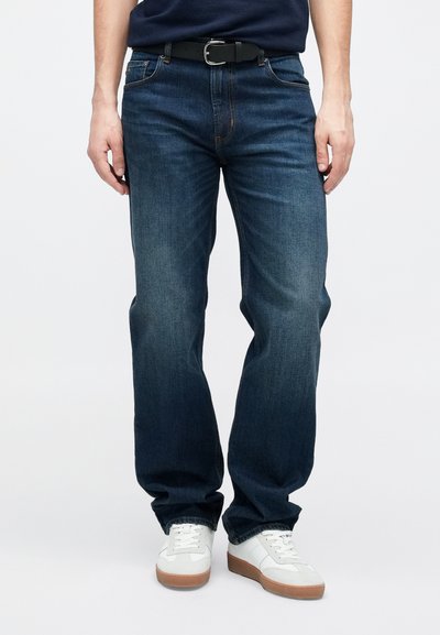 Σκούρο μπλε τζιν με ίσια γραμμή στα πόδια, με εμφανείς ραφές, πέντε τσέπες και φθαρμένες λεπτομέρειες. Συνδυασμένο με λευκά αθλητικά παπούτσια.