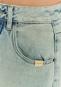 Světle modré džíny z denimu s jemně popraskanou texturou, přední kapsou s malou krémovou etiketou a kontrastním zlatým šitím.