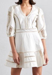 Robe en coton blanc avec un col en V, des manches bouffantes et des découpes en dentelle. Présente des rayures horizontales de garniture et un ourlet évasé.