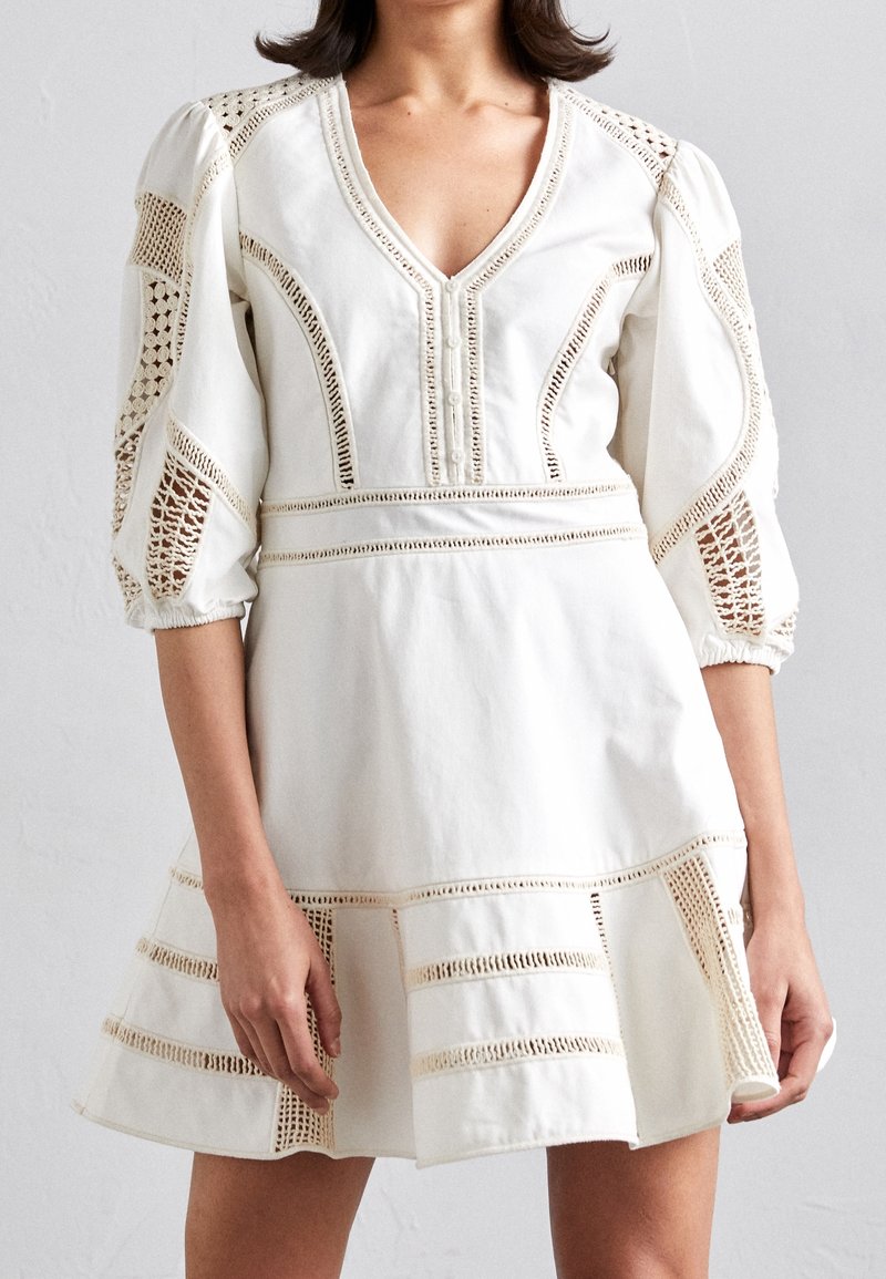 Robe en coton blanc avec un col en V, des manches bouffantes et des découpes en dentelle. Présente des rayures horizontales de garniture et un ourlet évasé.