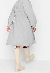 Grau, mittellange Oberbekleidung mit tailliertem Schnitt, weiten Ärmeln und seitlichen Taschen. Kombiniert mit knielangen beigen Gummistiefeln.