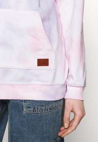 Jasnofioletowa i różowa bluza z kapturem w stylu tie-dye z przednią kieszenią, z brązową skórzaną naszywką z logo Roxy. Miękki materiał bawełniany.