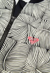 Tissu à motif de feuilles noir et blanc avec une fermeture éclair noire et un petit logo rose en forme de cœur près de la fermeture éclair.