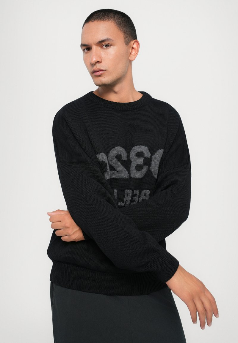 Pull en tricot noir avec un col rond, arborant un motif graphique gris contrastant sur la poitrine et des manches amples.