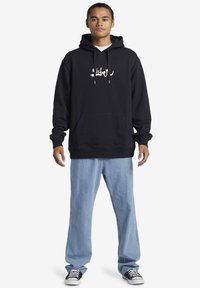 Sudadera negra con capucha y bolsillo frontal, estampado gráfico blanco, cordones, combinada con jeans de pierna ancha de color azul claro y zapatillas negras.