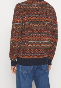 Man som bär en marinblå, orange och brun mönstrad tröja med geometriska mönster och blå denimjeans, sedd bakifrån.